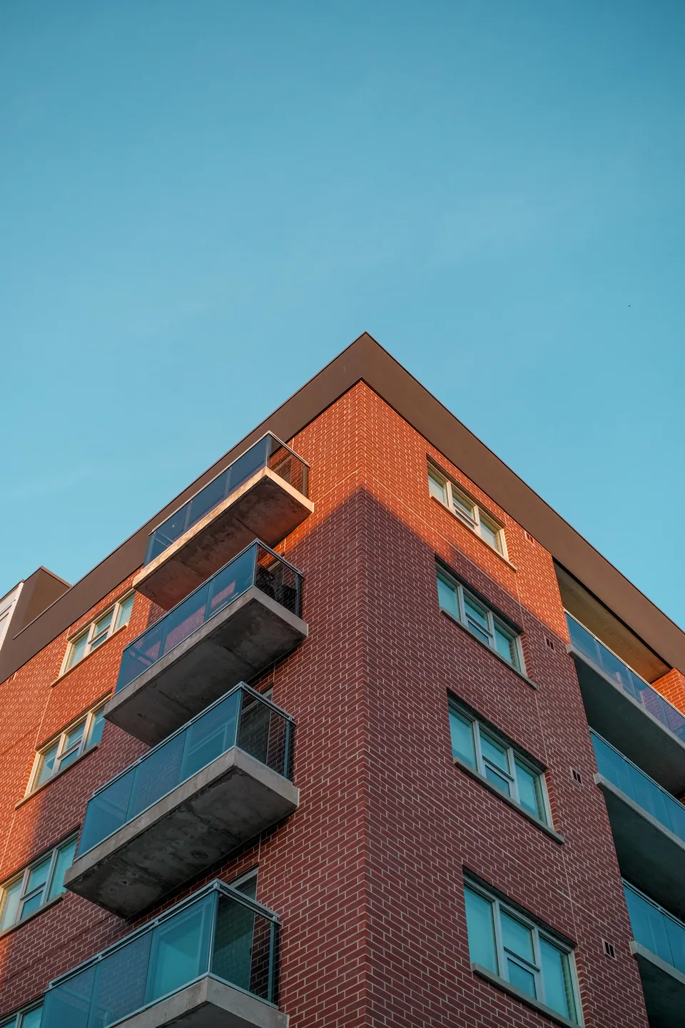 Illustrative photograph: Upper corner of a red brick residential building with projecting balconies against blue sky